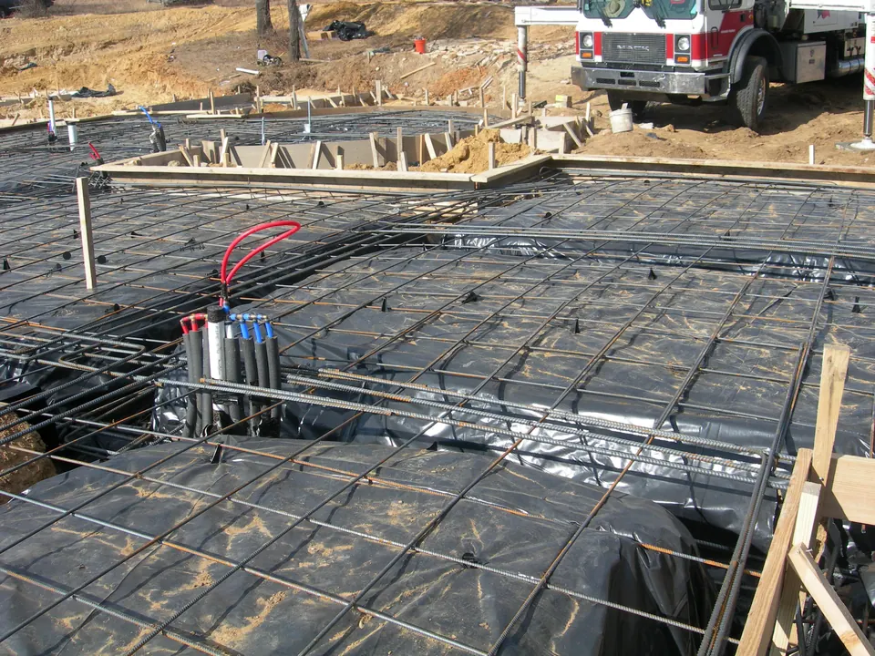 Rebar grid over vapor barrier inside foundation forms, with plumbing stubs and a concrete truck behind